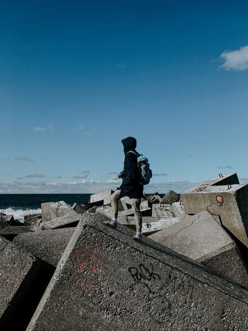 An individual standing on rocky concrete blocks by the sea under a clear blue sky.