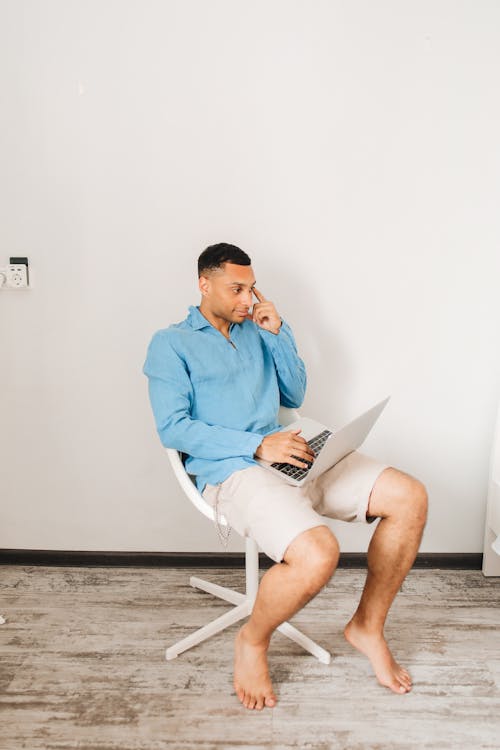 Free Man in Blue Long Sleeve Shirt Sitting on White Chair while Holding Silver Laptop Stock Photo