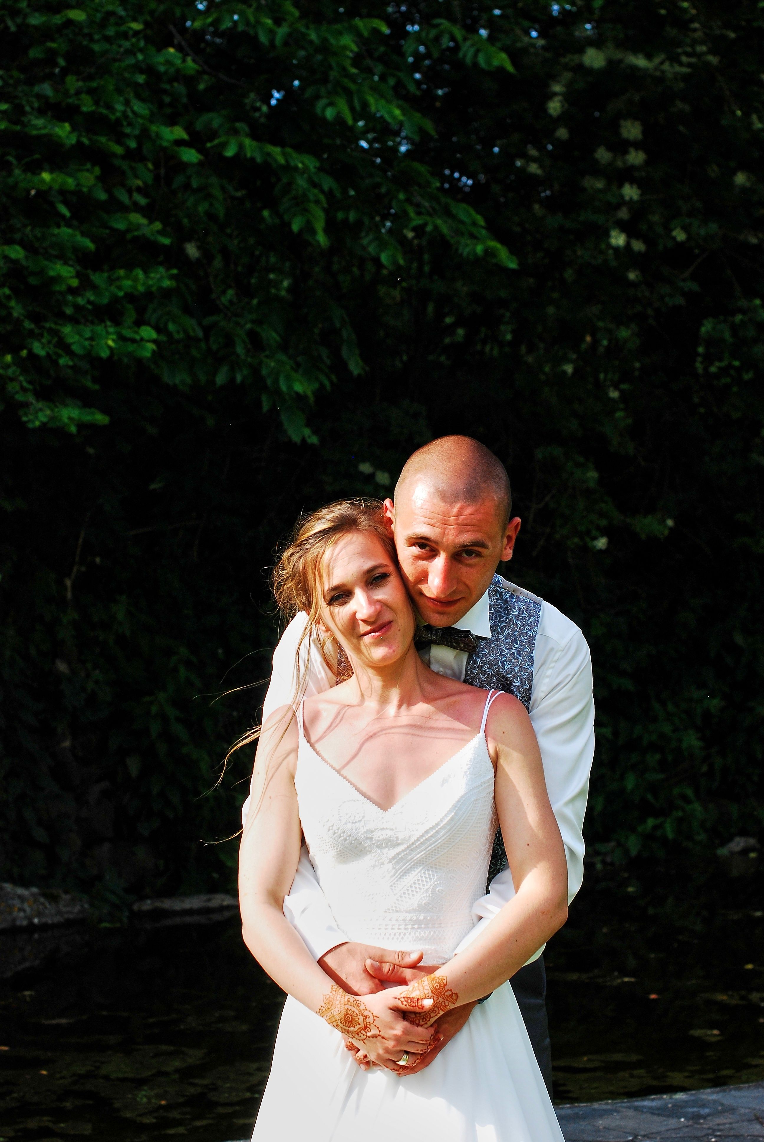 A loving couple embrace outdoors with green trees, the bride in a white dress and groom in a vest.
