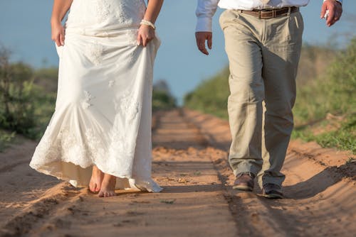 Free Woman in a Long White Dress and a Man in Brown Shoes Walking on Dirt Path Stock Photo
