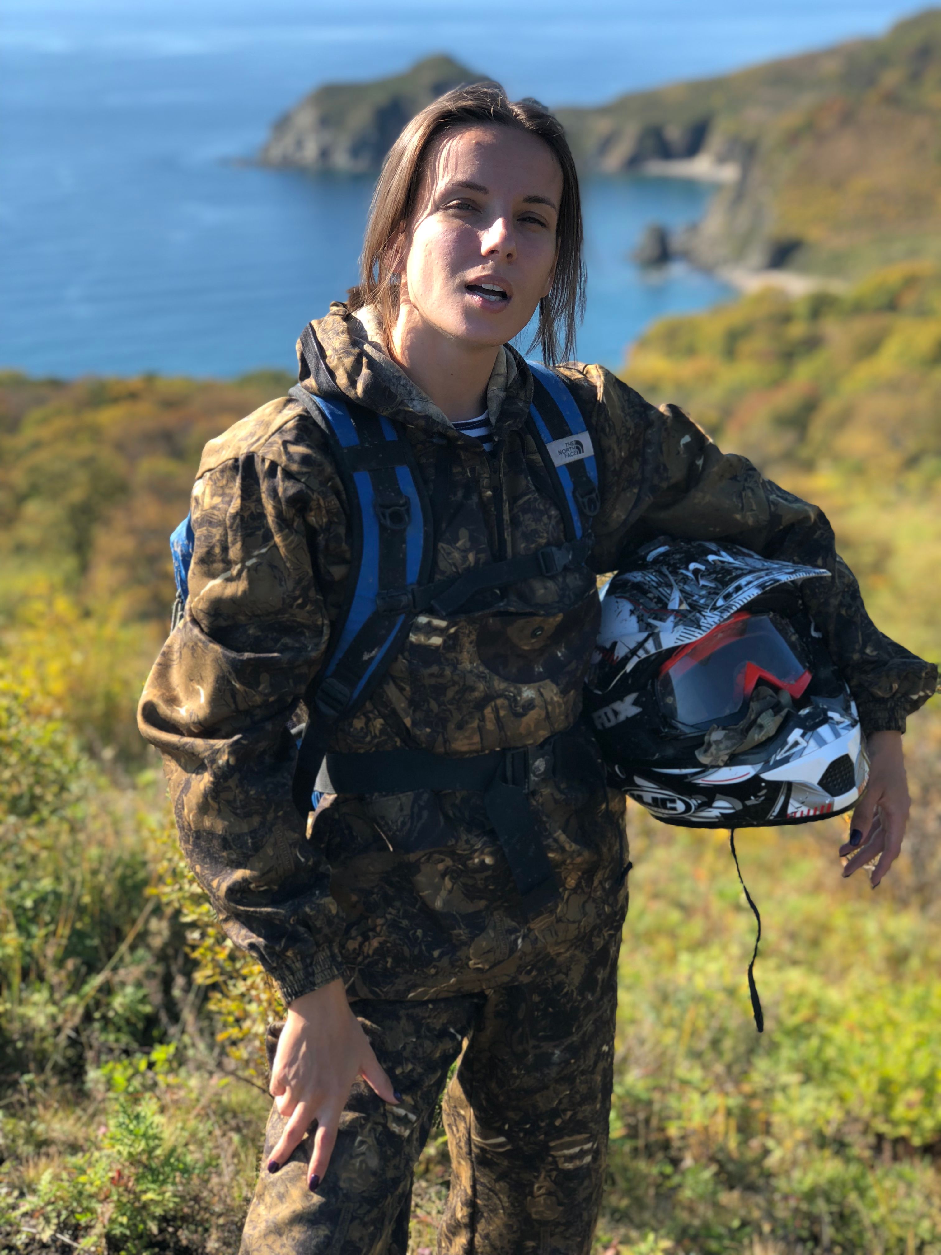 Woman in camouflage holding helmet, standing in scenic coastal area, Zato Fokino, Russia.