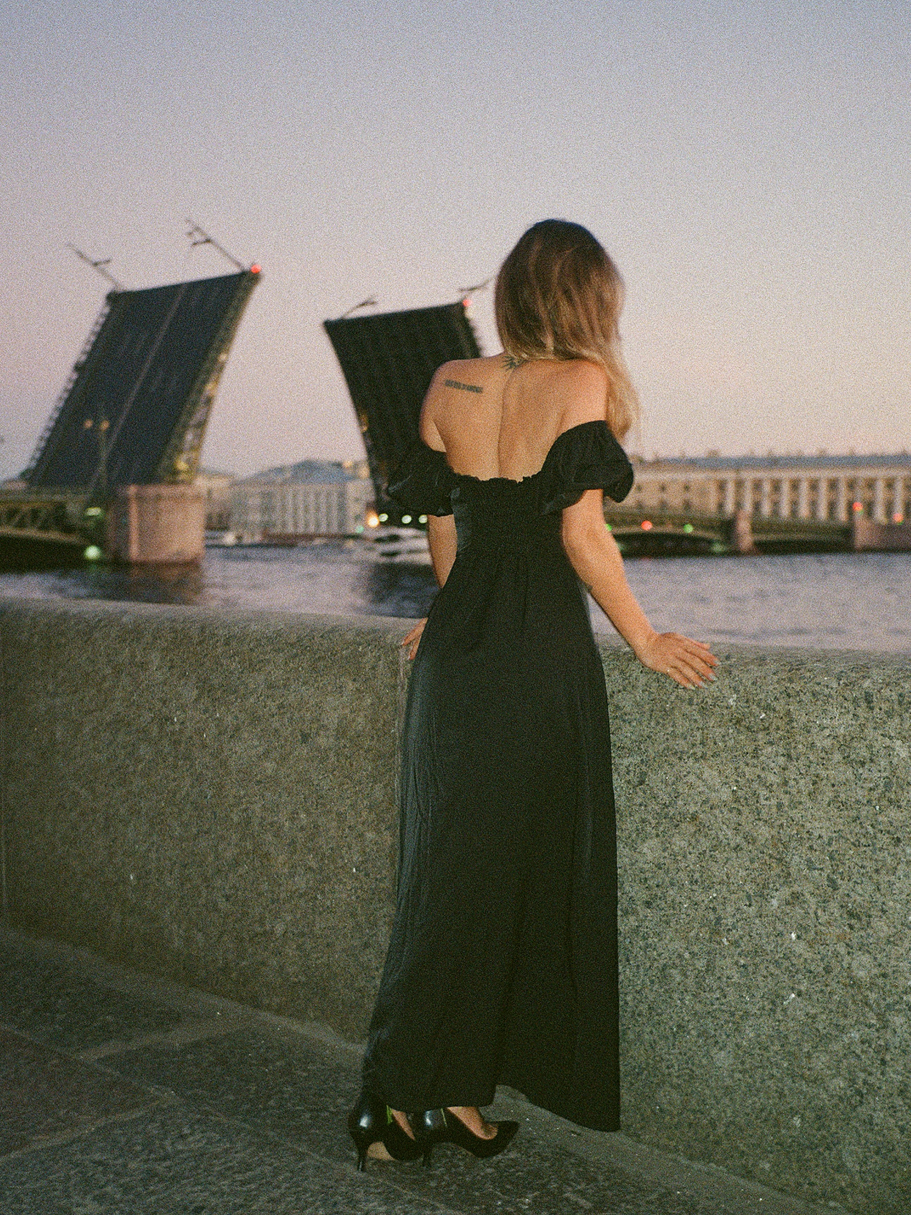 Woman in a black dress stands by a riverfront, gazing at an opened drawbridge during twilight.