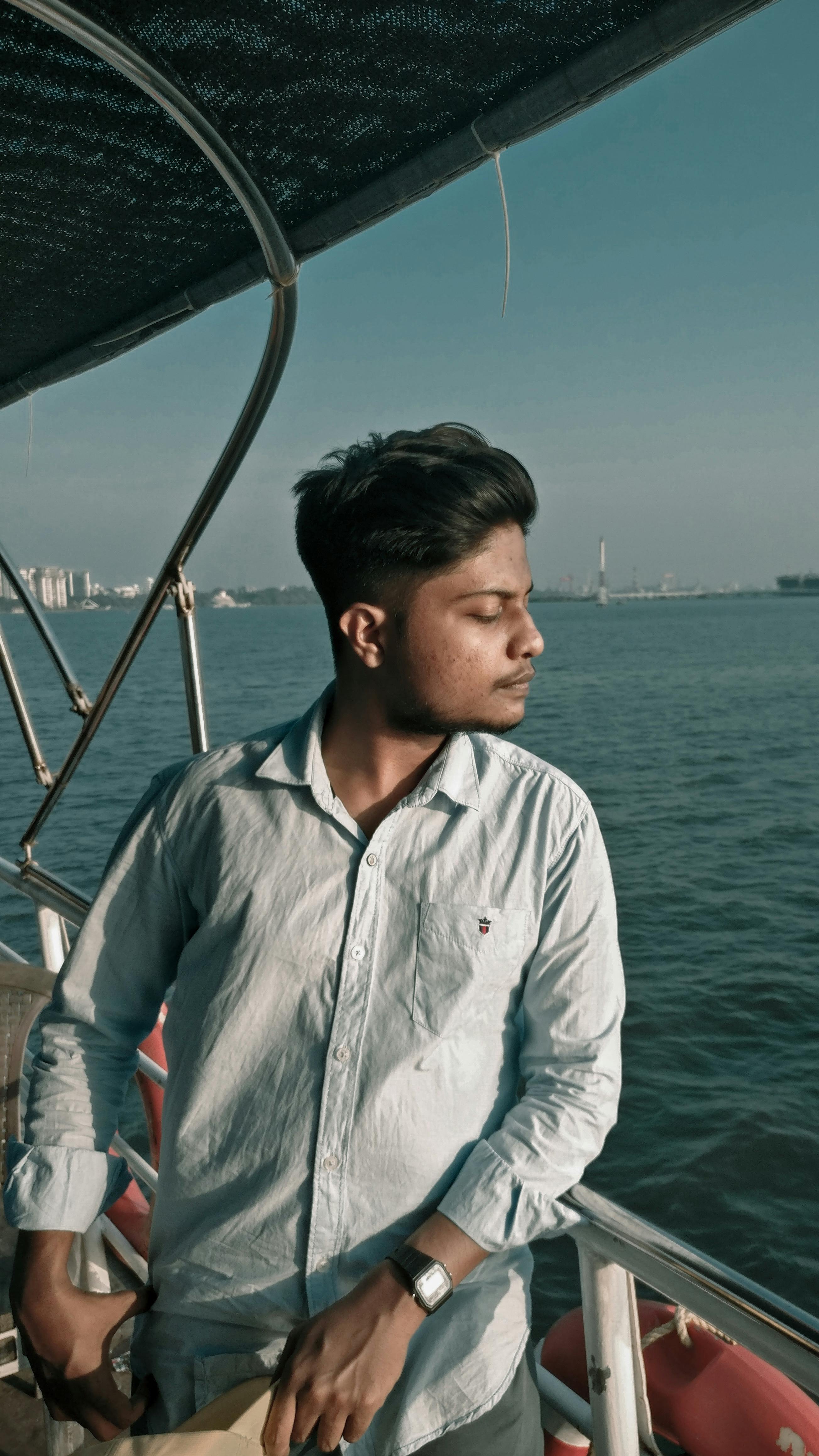 A young man stands on a boat with eyes closed, basking in sunlight by the ocean.