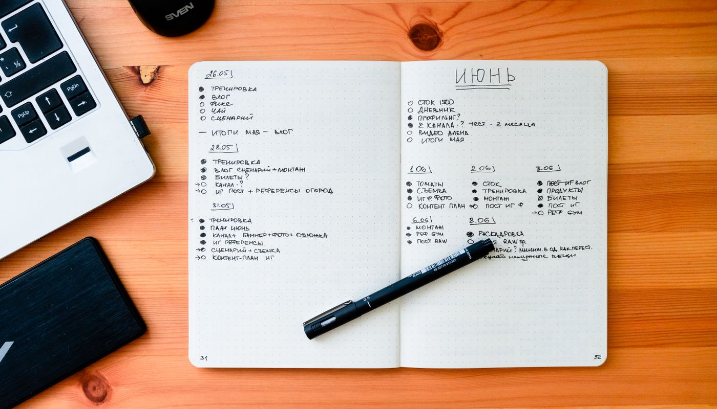 An open notebook with handwritten lists beside a laptop and pen on a wooden desk