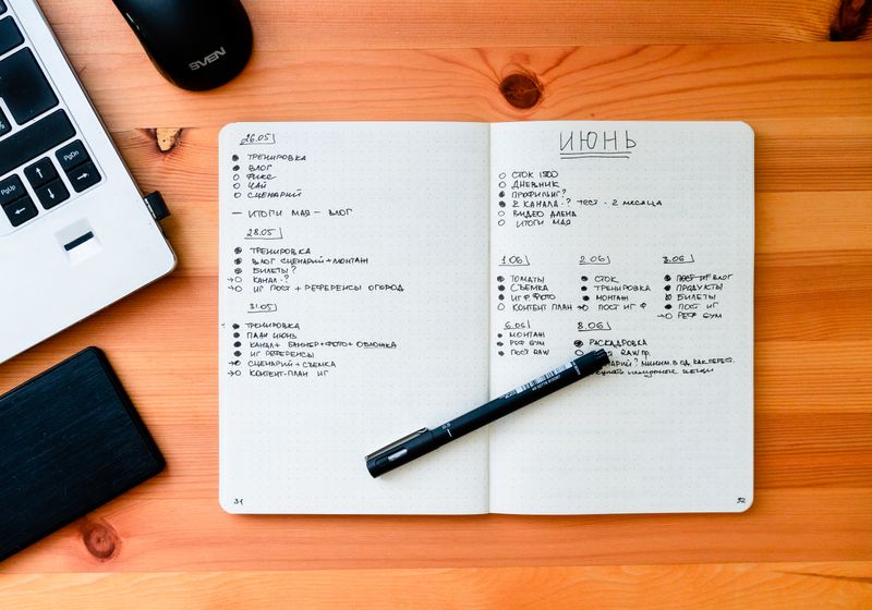 An open notebook with handwritten lists beside a laptop and pen on a wooden desk