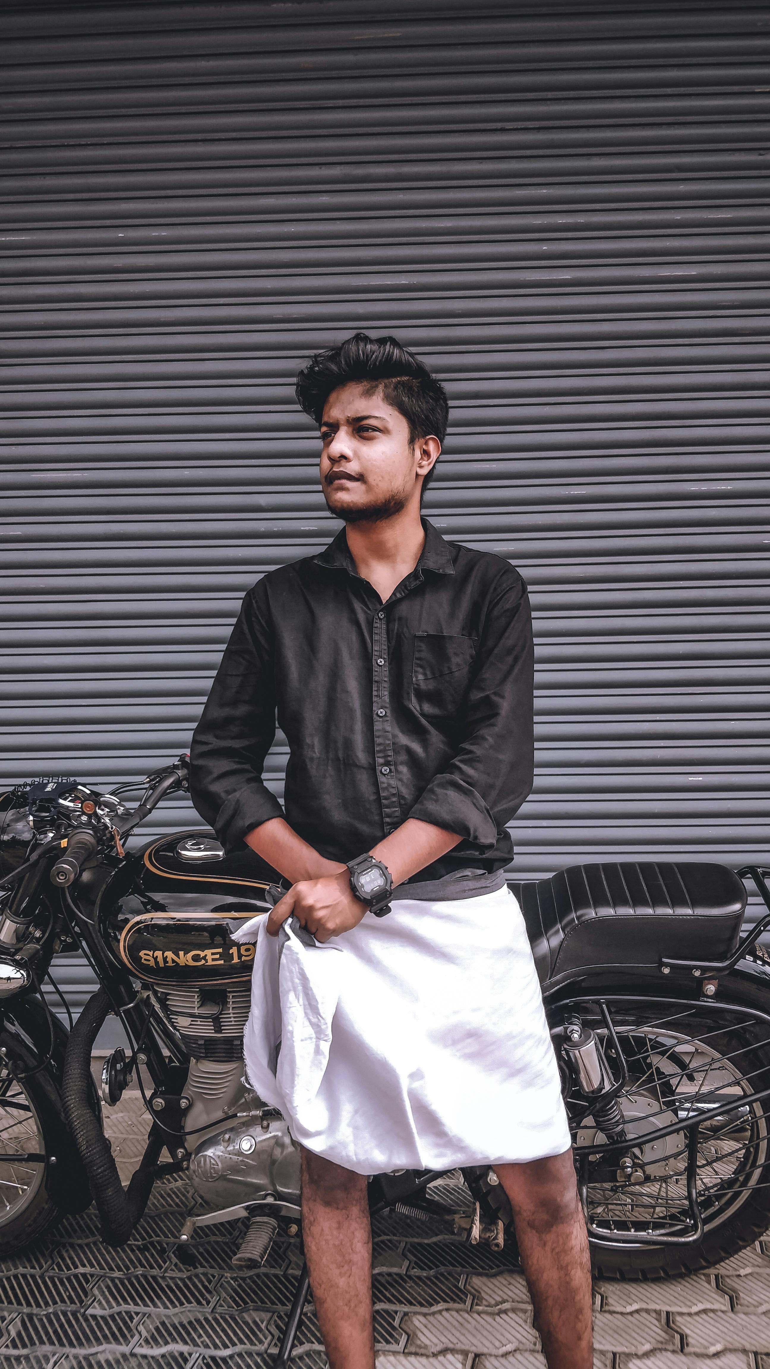 Young man in traditional attire posing confidently beside a vintage motorcycle.