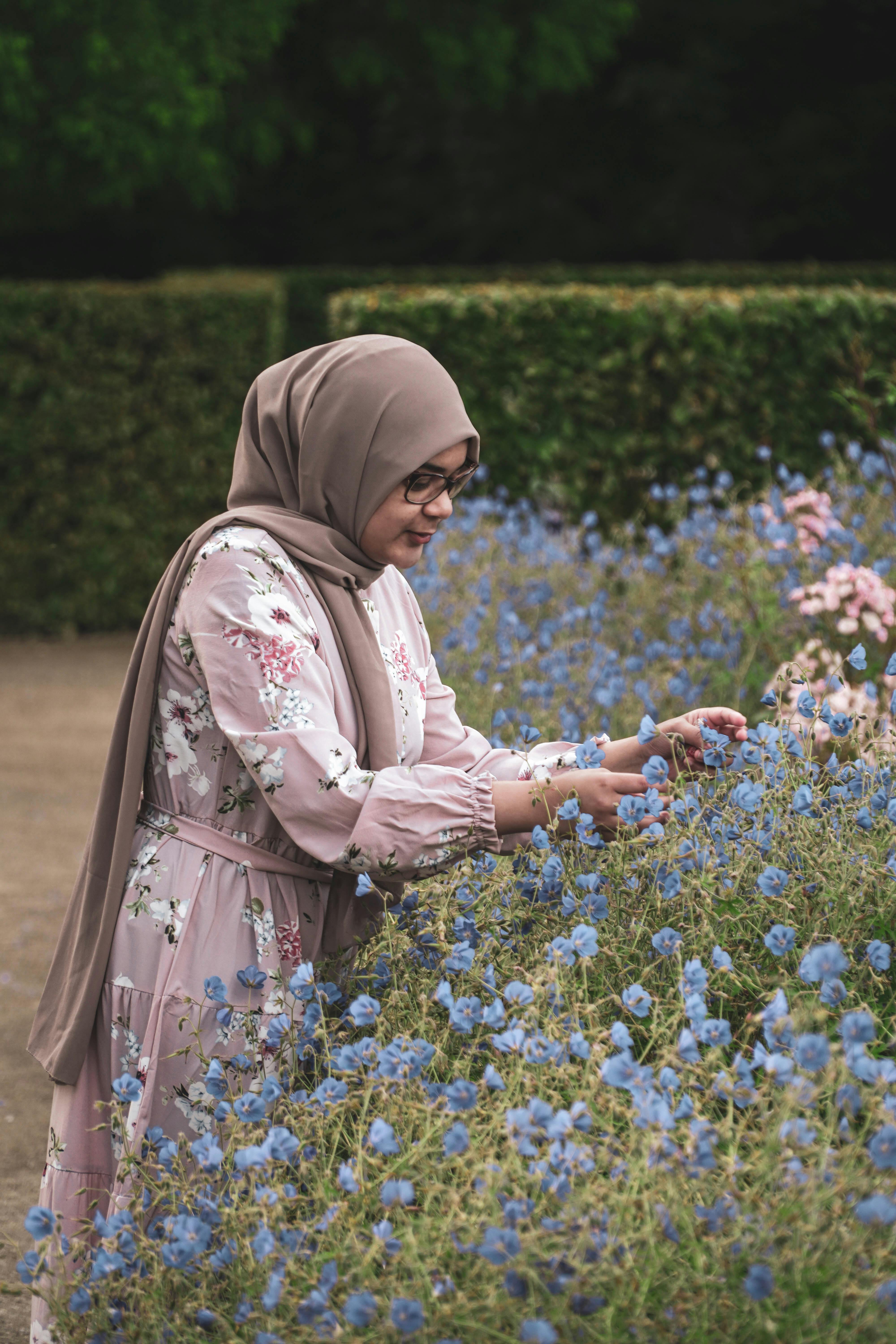 A woman in a floral dress and hijab admires blue flowers in an outdoor garden.