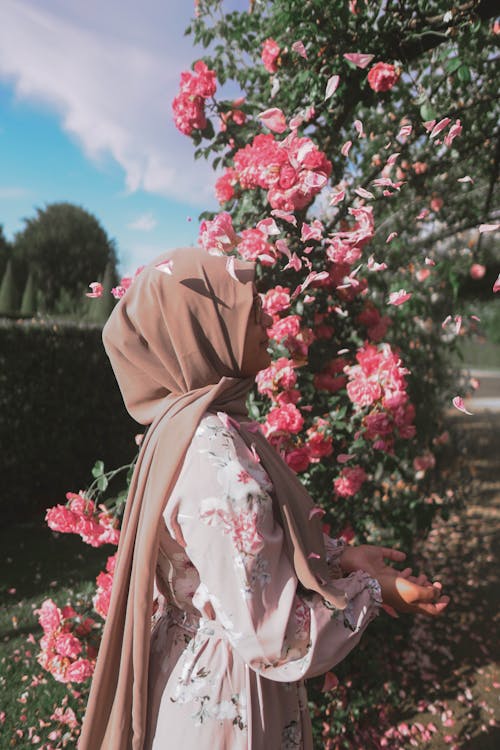 Side View of a Woman in Hijab and Floral Dress Standing near the Pink Flowers