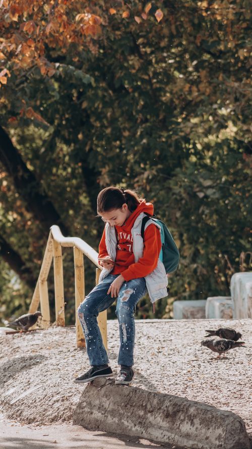 Free A young girl sitting outside looking at her cellphone surrounded by trees and birds. Stock Photo
