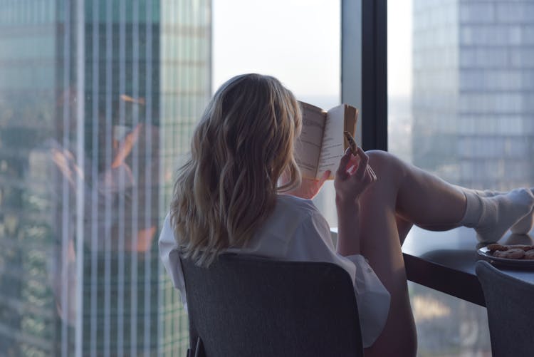 A Woman Reading A Book While Sitting On The Chair And Legs Up The Table