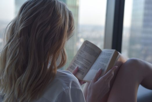 A woman is relaxing and reading a book by a large window with a city skyline view.. pexels, woman, relaxing