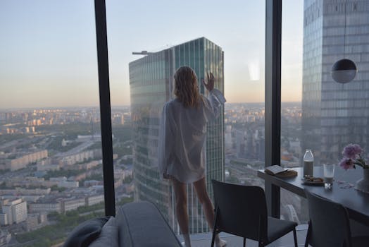 A woman in white stands by a window, overlooking a vast urban cityscape at dusk, conveying a sense of contemplation.
