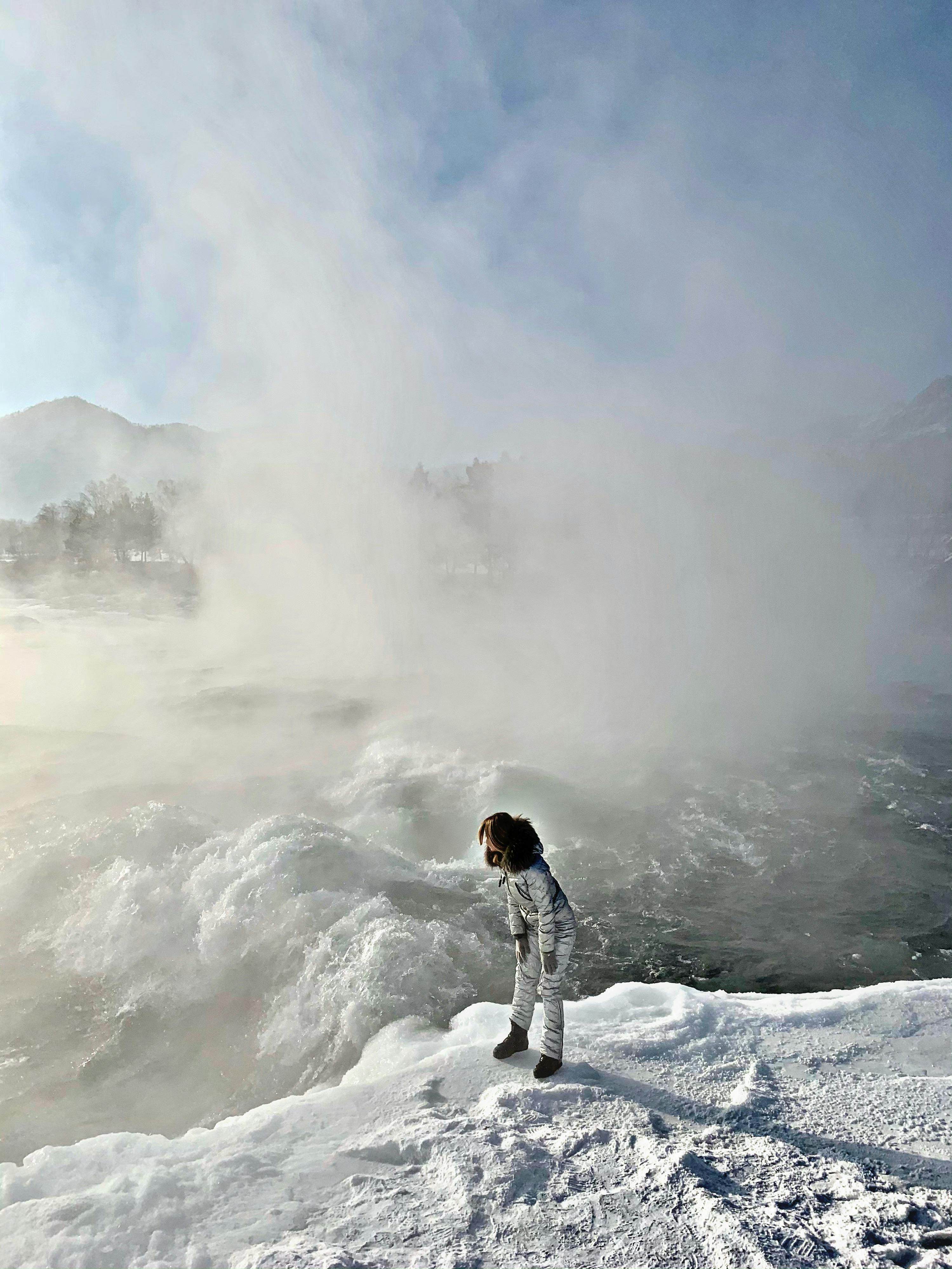 woman in winter overall standing on snow by a rough river and vapour in air