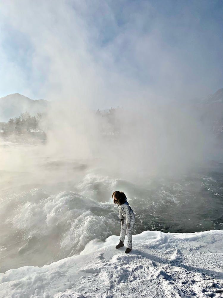 Woman In Winter Overall Standing On Snow By A Rough River And Vapour In Air