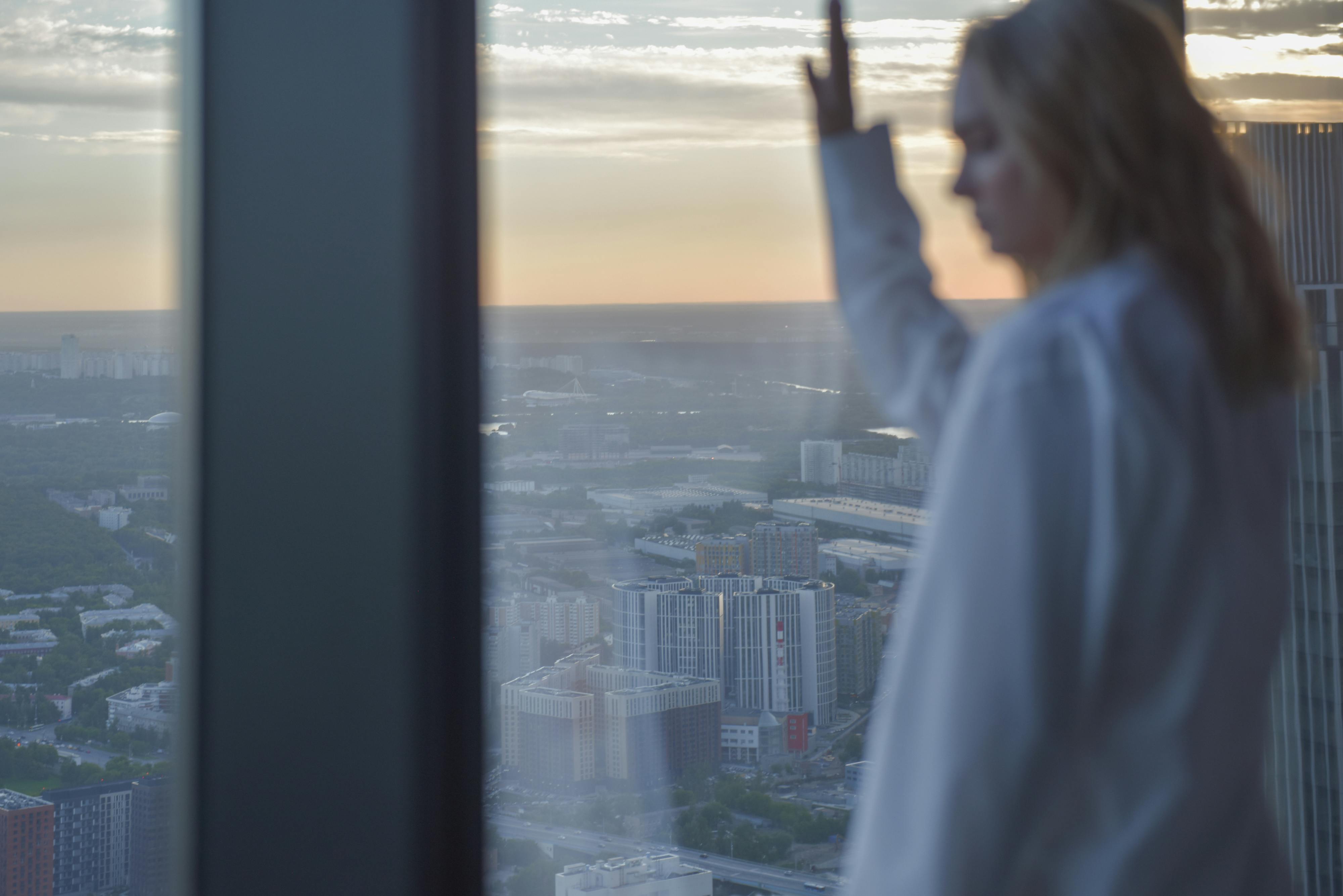 A pensive woman gazes out of a high-rise window over a cityscape at sunset.