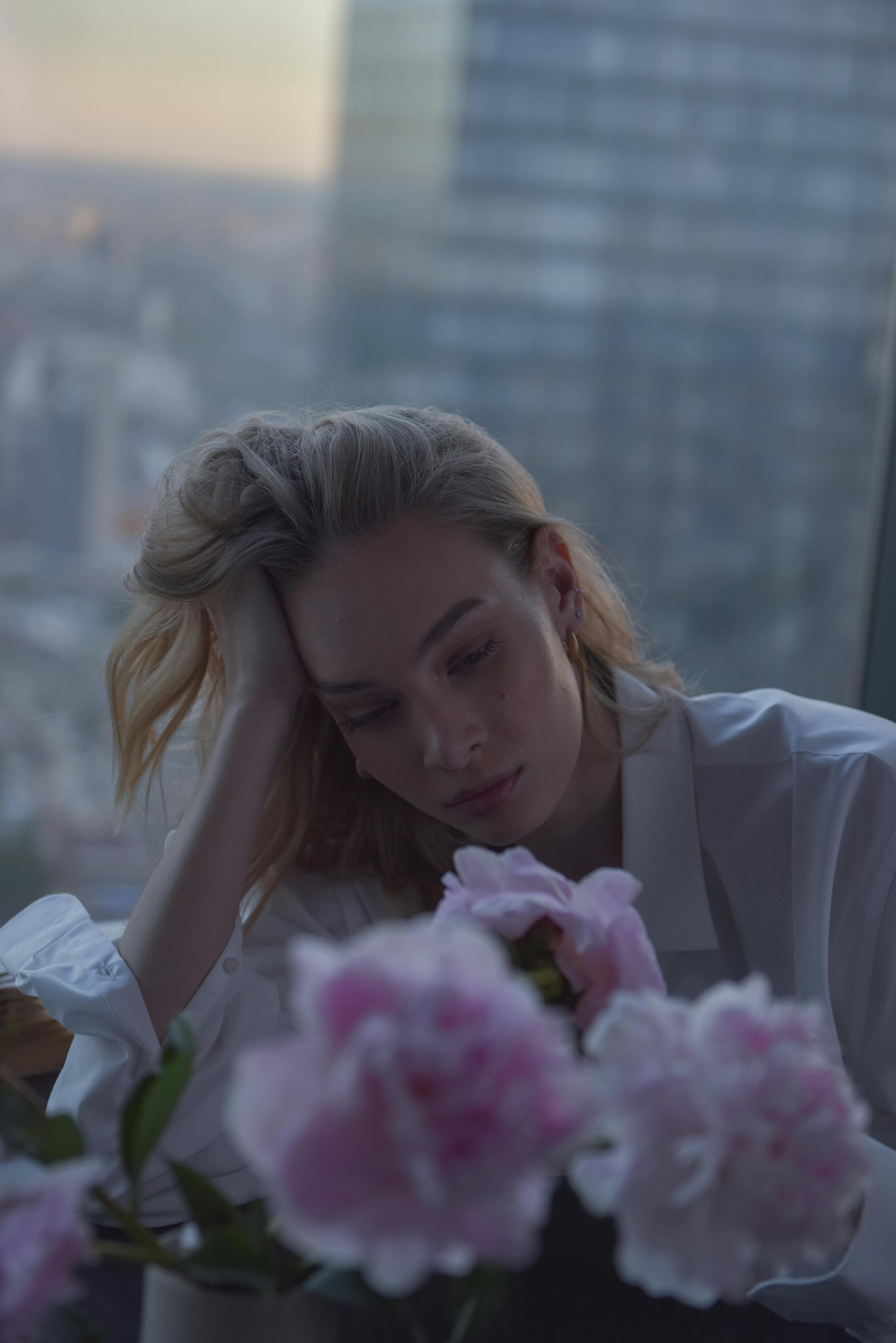 A thoughtful woman gazes at pink flowers indoors during sunset, in an urban setting.