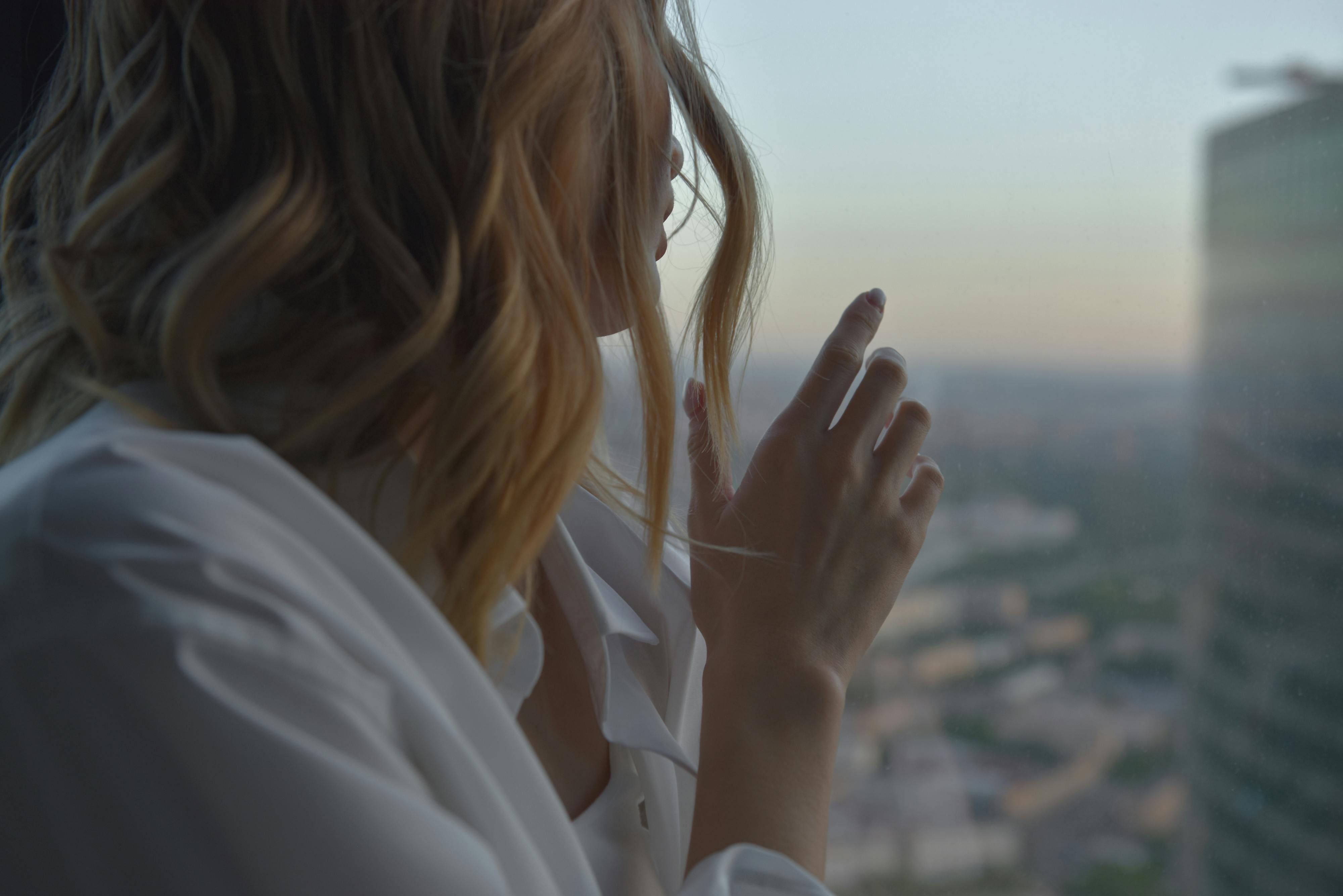 Woman in a white shirt looking out a window at dawn, capturing a moment of reflection.