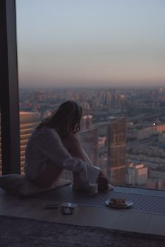 A woman sits by a high-rise window, reflecting on the cityscape at dusk.