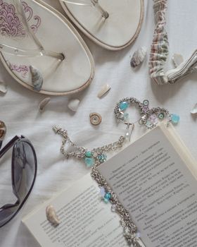 Stylish beach essentials including flip-flops, jewelry, sunglasses, and an open book on white fabric.