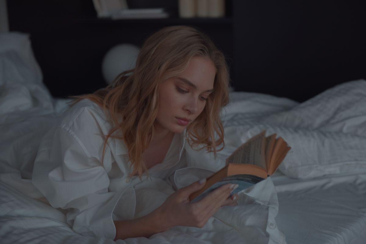 A woman lying comfortably on a bed reading a book with a calm expression in a cozy bedroom setting.