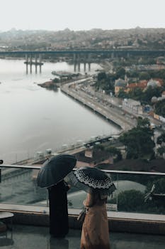 People holding umbrellas overlook a scenic urban riverside view under a cloudy rainy sky.