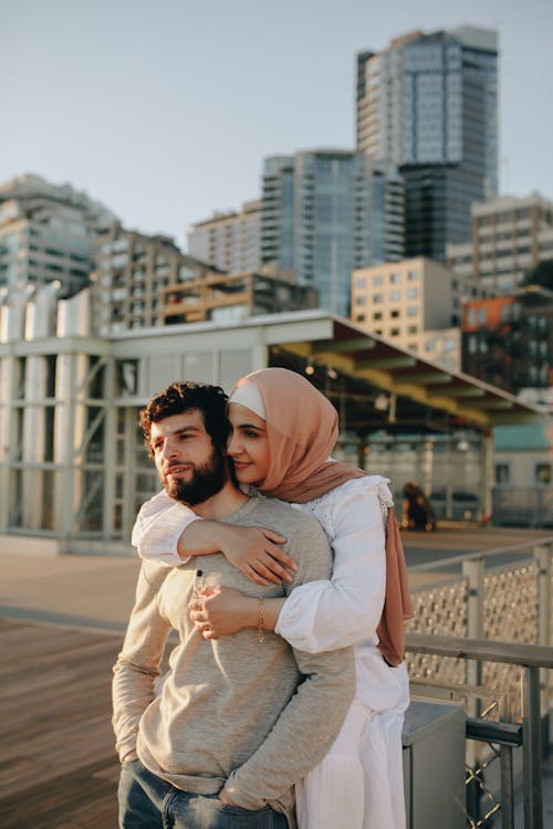 Free Woman Wearing Headscarf Embracing Bearded Man and Modern Architecture in Background Stock Photo