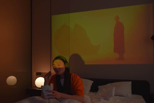 An Asian woman with headphones enjoys eating on bed watching a movie projection.