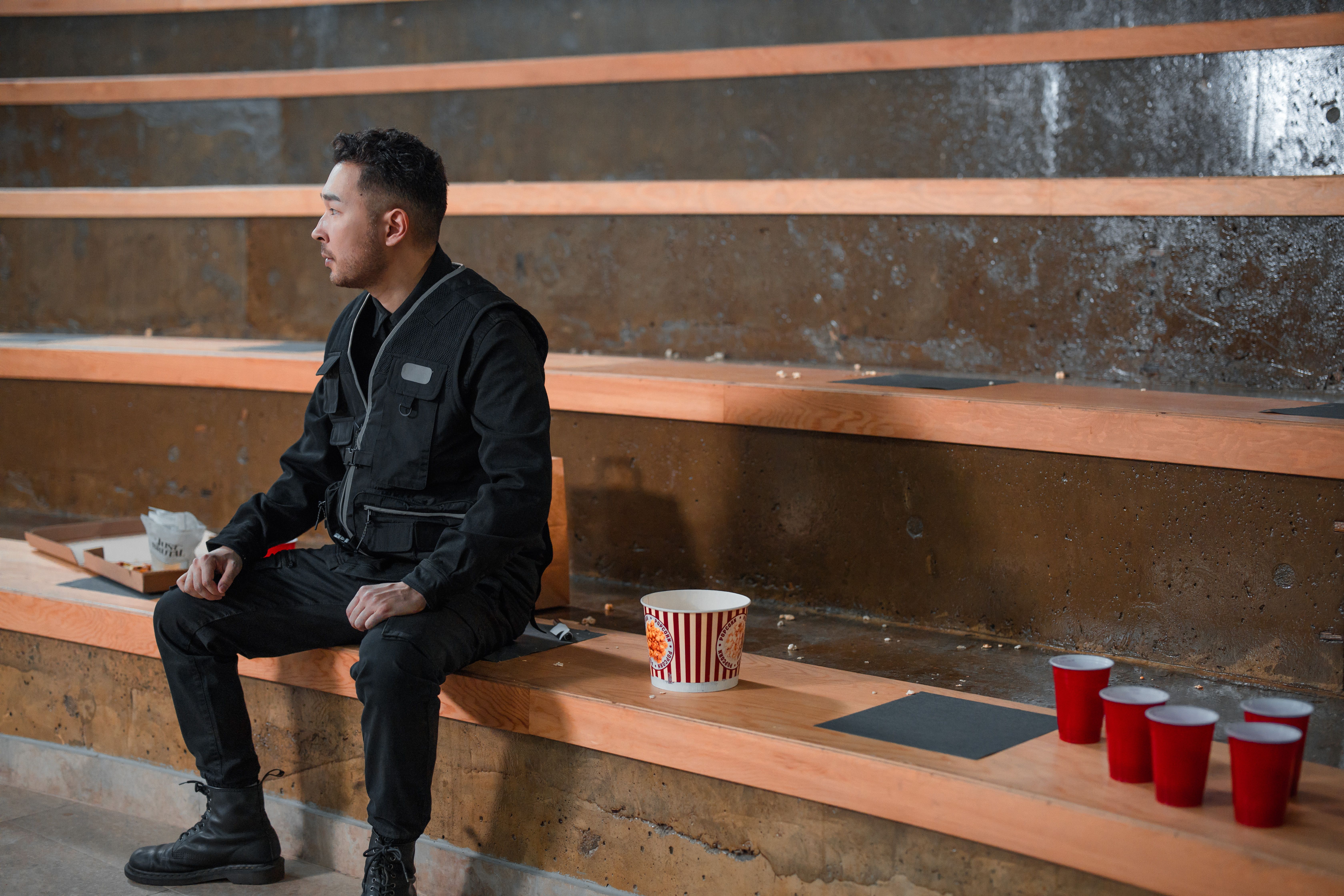 A man in black uniform sits alone on wooden steps surrounded by empty snack containers and cups.
