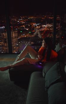 Woman with long hair relaxing on a sofa, enjoying a city skyline view at night.