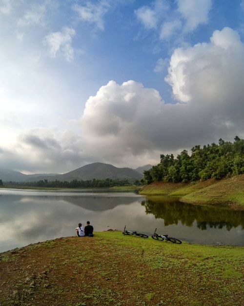 Free A couple sits by a tranquil lake with bikes, enjoying the lush landscape and serene atmosphere. Stock Photo