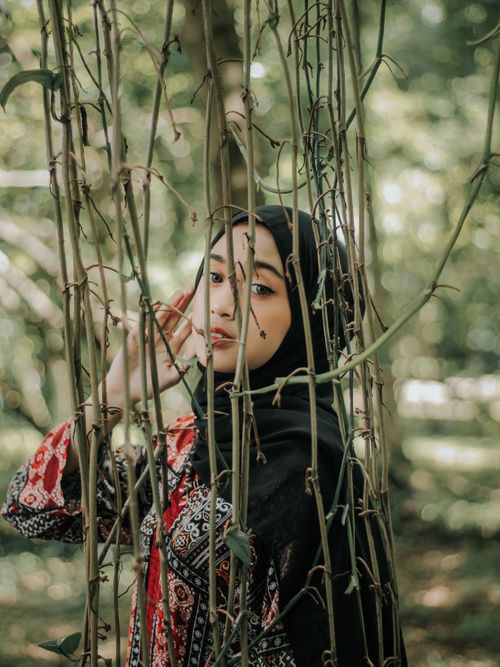 Free A woman in a red patterned dress and hijab elegantly poses among branches, captured in a serene outdoor setting. Stock Photo