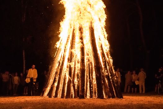 A large bonfire illuminates the night during a festive gathering in Utena, Lithuania with people watching attentively.