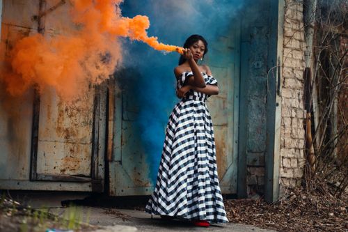 Free A stylish woman in a checkered dress poses with vibrant smoke outdoors against a rustic backdrop. Stock Photo