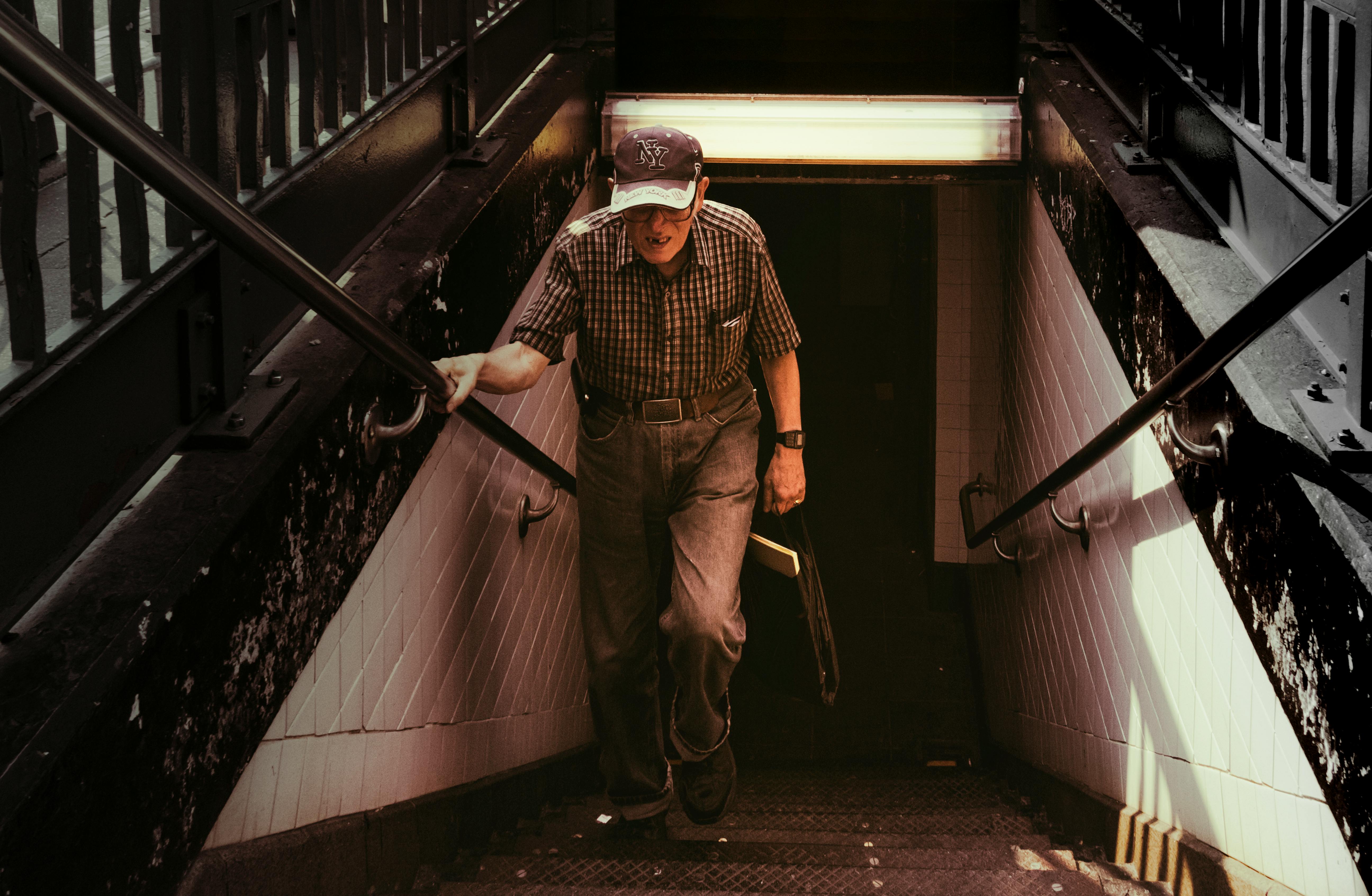 Senior man climbing subway steps in New York City, wearing a cap and carrying a newspaper.
