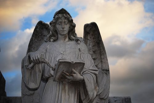 Free Serene stone angel sculpture holding an open book against a dramatic sky backdrop. Stock Photo