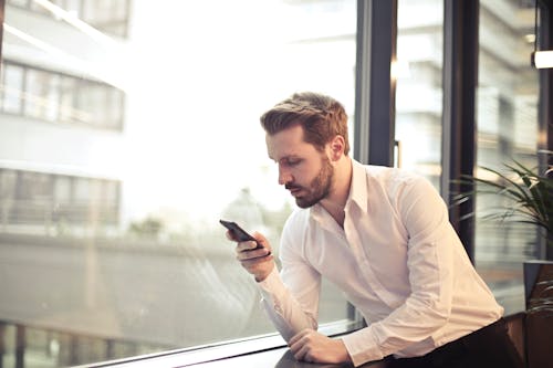 Zdjęcie Mężczyzny W Białej Koszuli Trzymającego Telefon W Pobliżu Okna