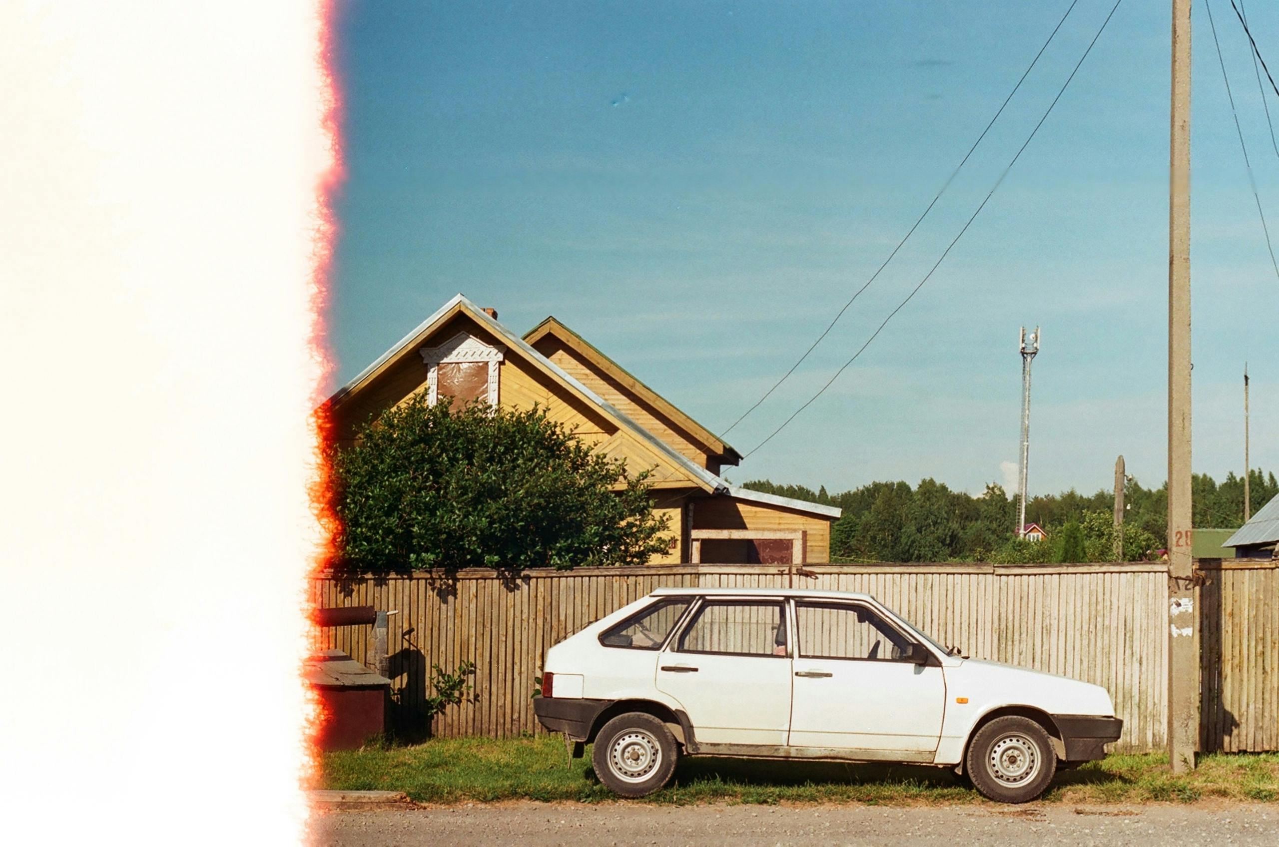 A vintage car parked near a rustic house under a bright blue sky on a countryside roadside.
