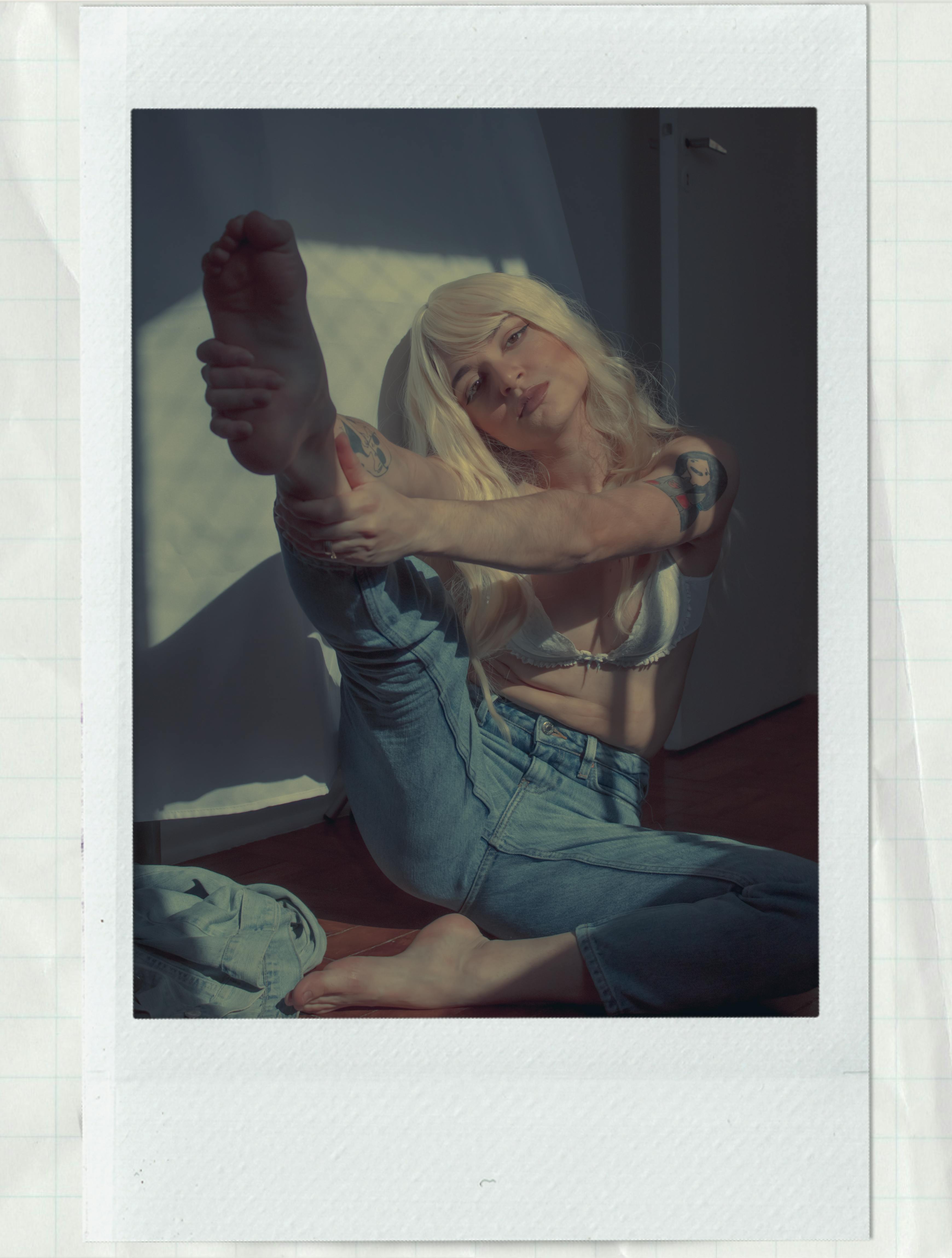 Blonde woman in blue jeans and white bra striking a pose indoors with natural lighting.