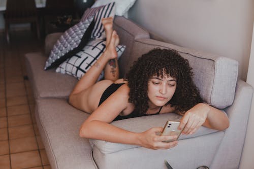 Woman with Curly Hair Lying on a Couch while Using Her Cell Phone