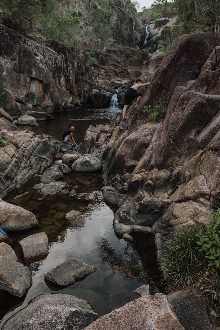 Man Sitting On Rocks In Middle Of Creek Running Through Mountains