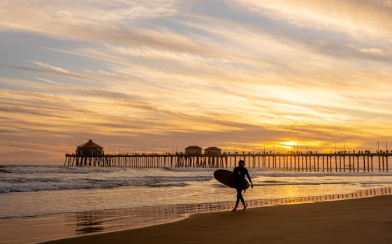 A surfer walks along Huntington Beach at sunset with a vibrant sky in the background.