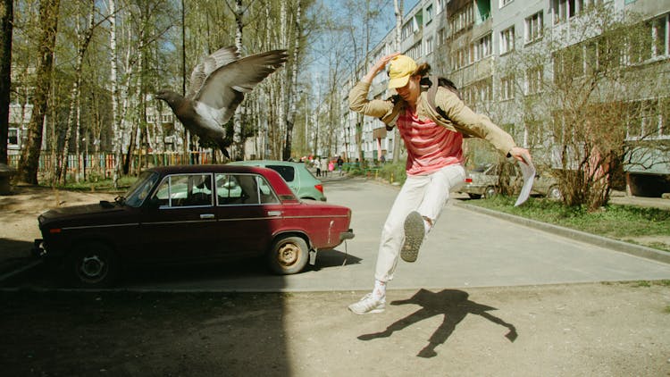 Man In White Pants And Brown Jacket Jumping Near A Bird And Car