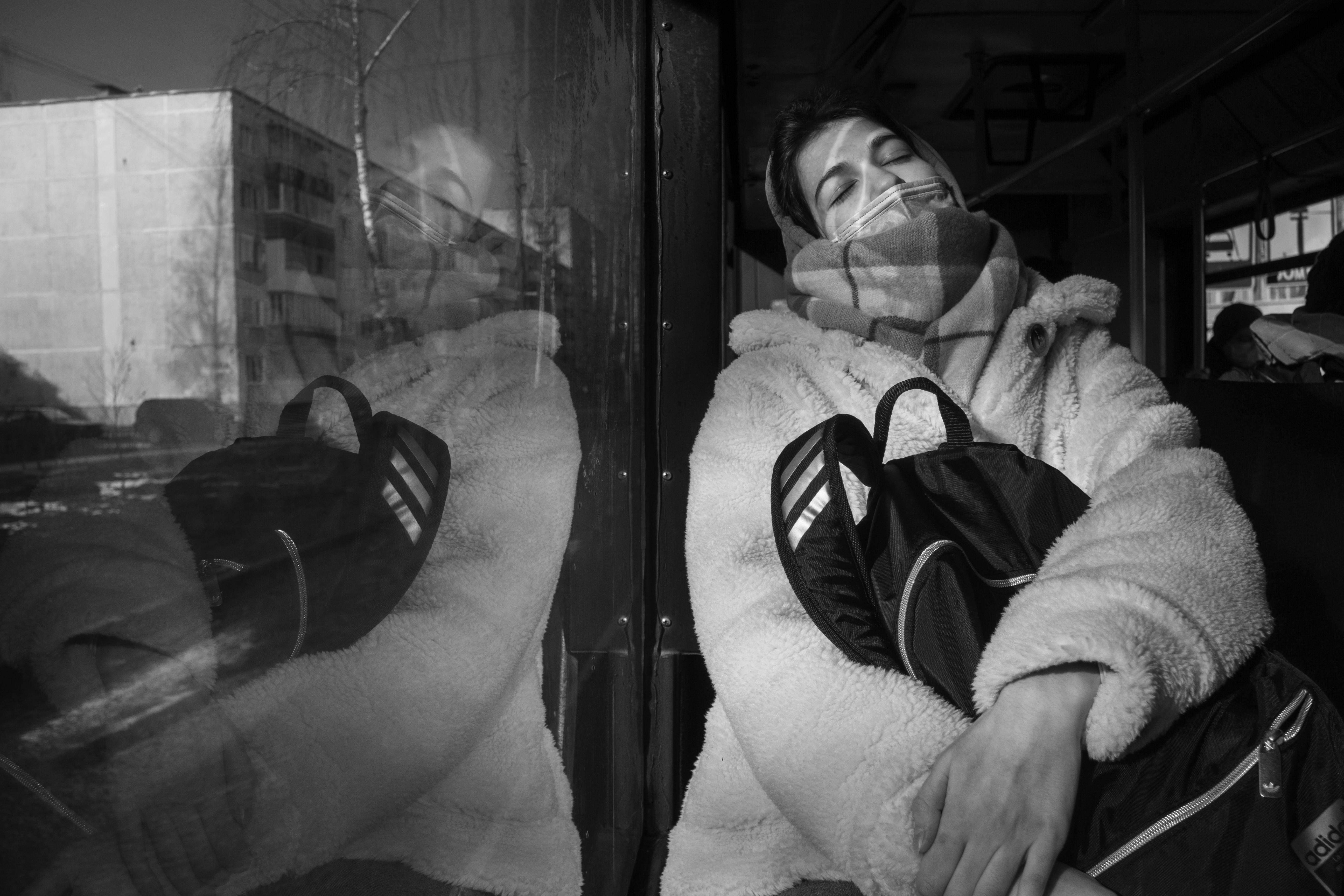 Black and white image of a person sleeping on a bus in Псков, Russia. Reflective glass shows urban scenery.