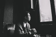 Grayscale Photo of Man Sitting Near Closed Window