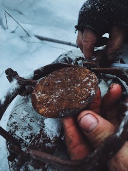 Hands wearing gloves hold a rusty metal object on snow-covered ground in a winter setting.