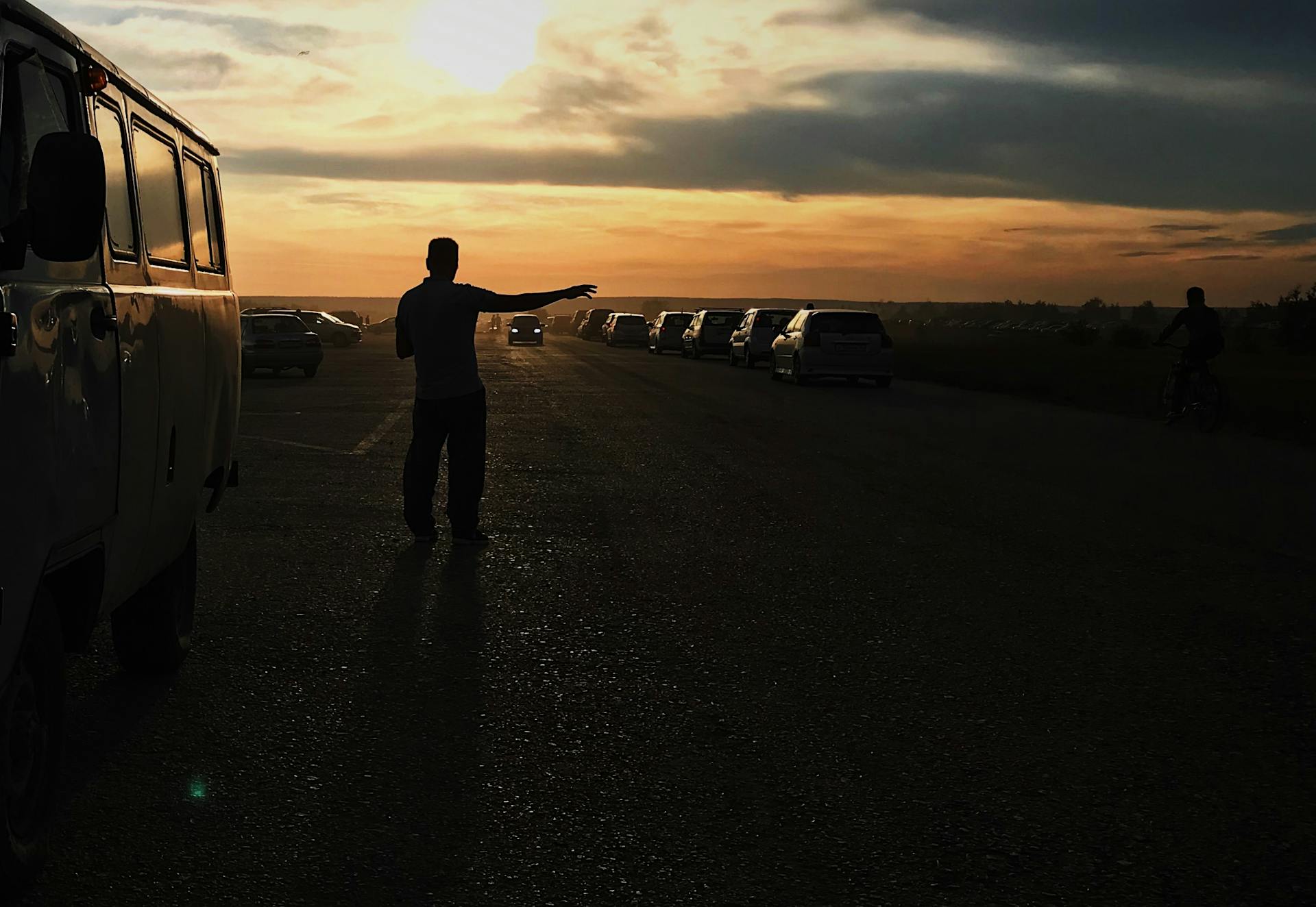 Silhouette of a person at a sunset-lit parking lot with lined cars and a cloudy sky.