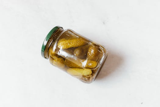 Glass jar with pickled cucumbers on a white surface, showcasing classic still life photography.