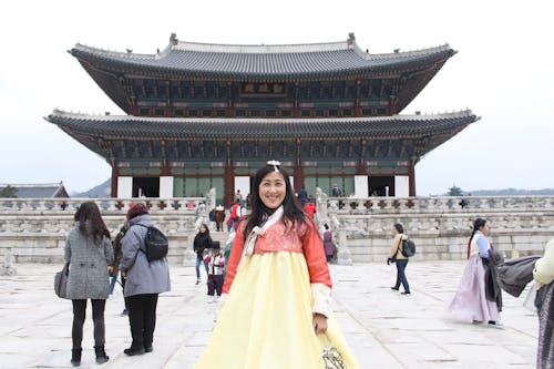 Positive Asian female tourist wearing long yellow dress standing on square in front of pagoda during vacation