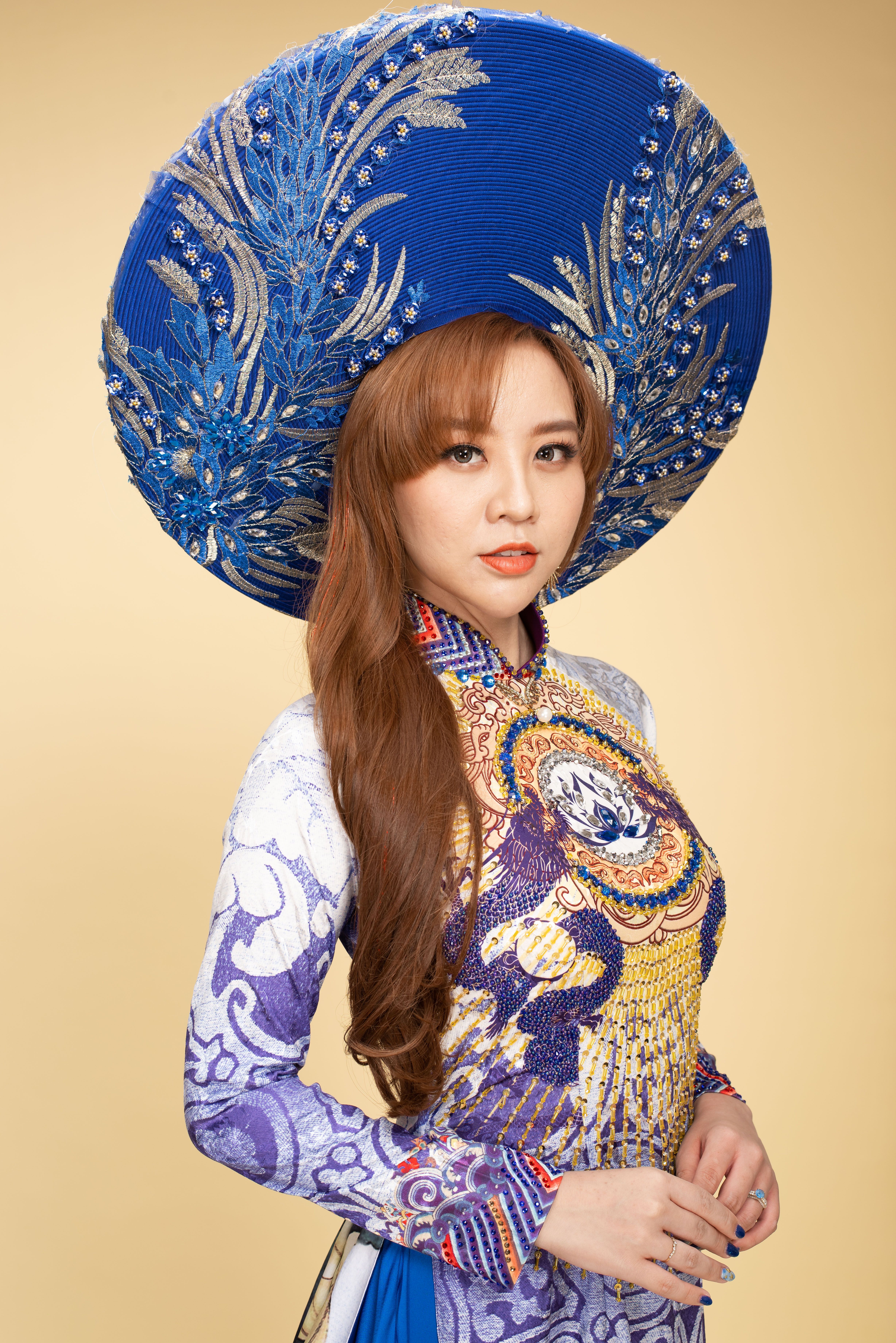 Portrait of an elegant Asian woman wearing a traditional dress and large blue hat in a studio setting.