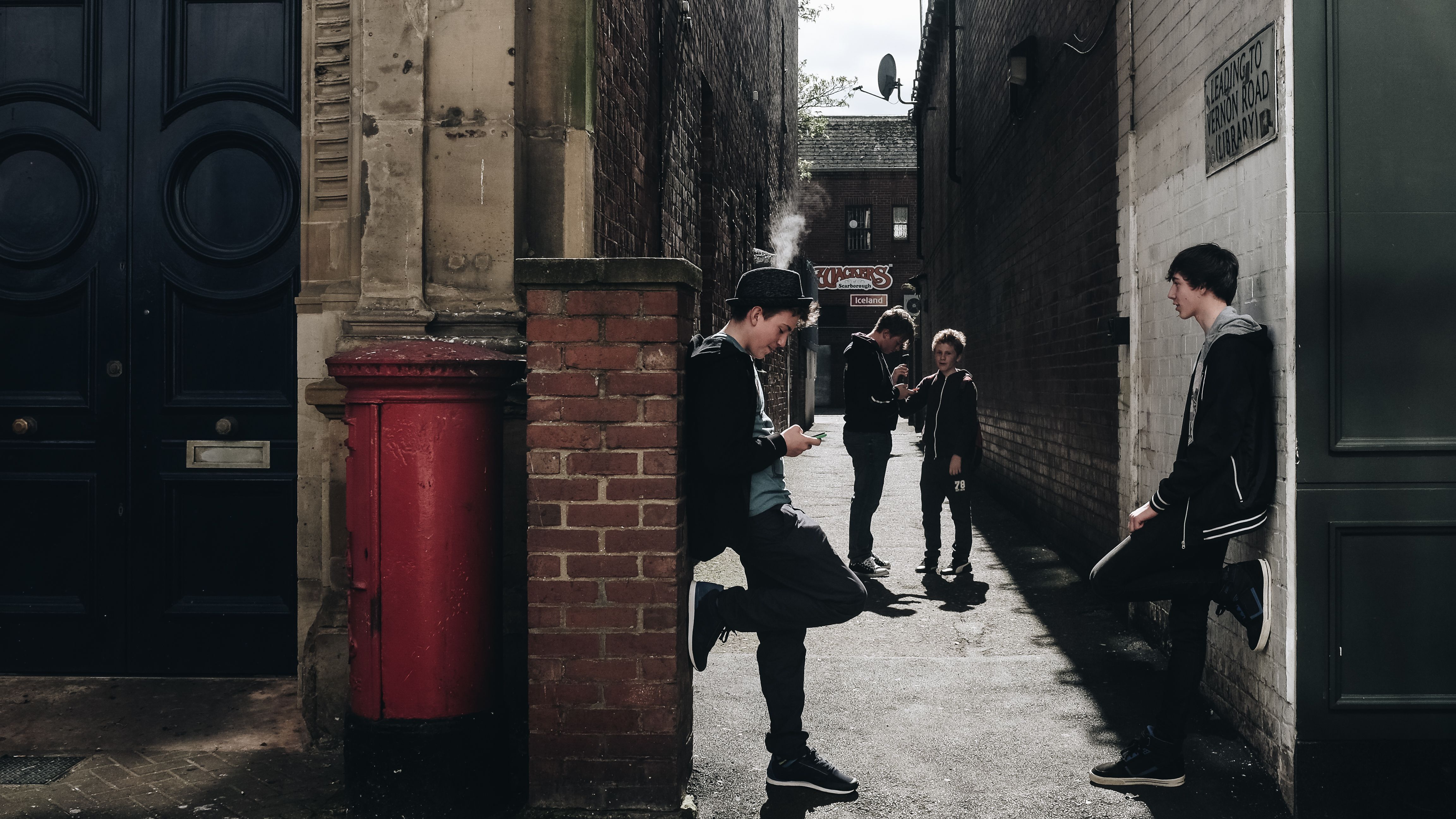 A group of teenagers socializing and leaning against walls in a narrow urban alley during the day.